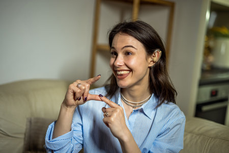 A joyful young woman sits in her living room wearing a hearing device, communicating confidently through sign language.の写真素材