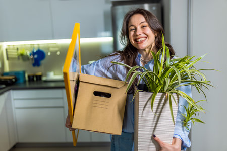 A cheerful young woman is entering her new apartment, holding boxes, a potted plant, and luggage.の写真素材