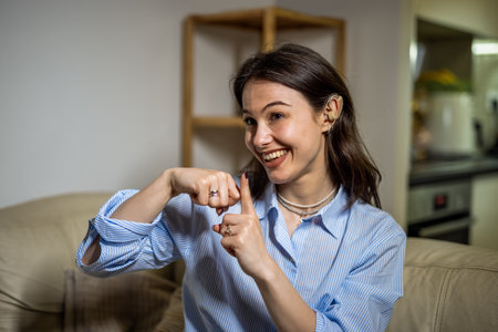 A joyful young woman sits in her living room wearing a hearing device, communicating confidently through sign language.の写真素材
