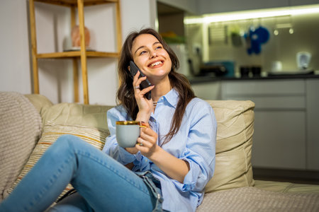 A young woman is sitting in her cozy living room, enjoying a cup of coffee while having a phone conversation.の写真素材