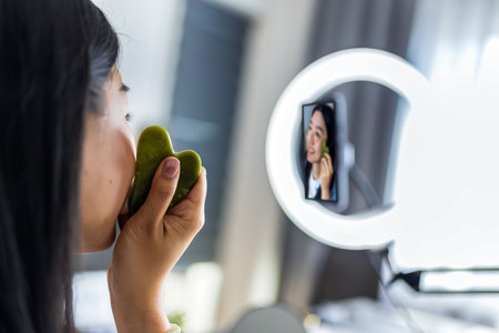 A young Thai influencer demonstrates her gua sha facial massage routine while filming a skincare video with a ring light.の写真素材
