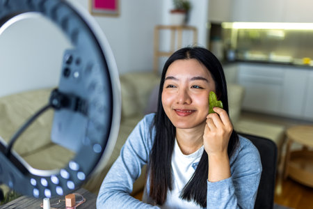 A young Thai influencer demonstrates her gua sha facial massage routine while filming a skincare video with a ring light.の写真素材