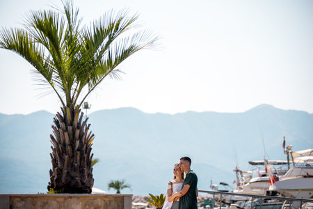 A young couple stands closely together on a marina deck, holding on to each other while admiring a tall palm tree.の写真素材