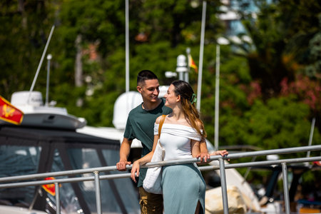 A young couple stands on a marina bridge with the boyfriend hugging his girlfriend from behind, both gazing peacefully at the sea and each otherの写真素材