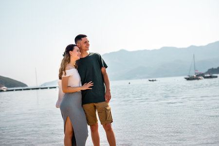 Young couple stands on the beach gazing into the distance, with the girlfriend resting her head gently on her boyfriend's chest.の写真素材