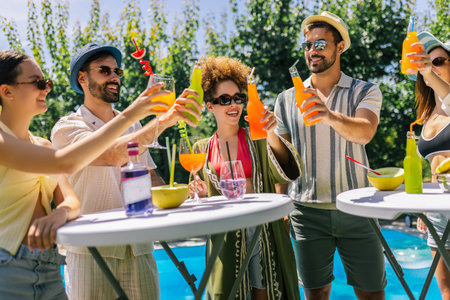 A joyful group of young people by the pool cheers with colorful cocktails, wearing swimsuits, hats,...