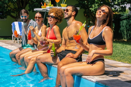 A group of young people in bathing suits sit by the pool with their legs in the water, enjoying...