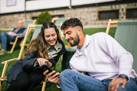 A young man and woman unwind on lounge chairs at a scenic mountain resort during a weekend getaway. They share a moment together while looking at a smartphone screenの写真素材