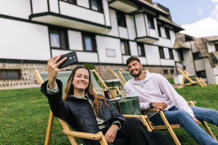 A young man and woman enjoy a weekend getaway at a scenic mountain resort. Lying back on lounge chairs, they capture the moment by taking a cheerful selfie with a smartphone.の写真素材
