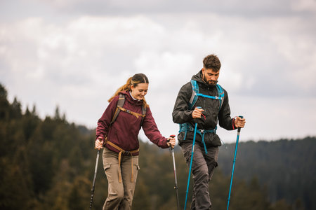 A young man and woman, fully equipped with backpacks, hiking poles, and outdoor clothing, walk together along a mountain trail surrounded by lush natureの写真素材