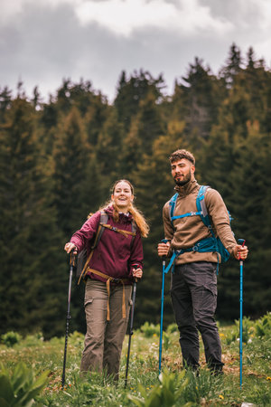 A young man and woman, fully equipped with backpacks, hiking poles, and outdoor clothing, walk together along a mountain trail surrounded by lush nature. They pause to admire the scenic beauty around them.の写真素材