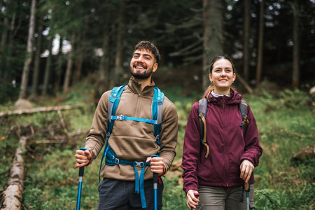 A young man and woman are captured hiking through a lush mountain forest, fully equipped with backpacks, hiking poles, and outdoor clothing. The couple appears focused and determined as they navigate the woodland trail.の写真素材