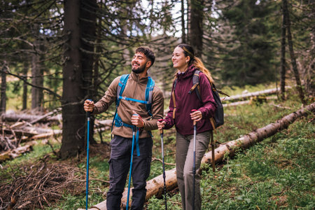 A young man and woman are captured hiking through a lush mountain forest, fully equipped with backpacks, hiking poles, and outdoor clothing. The couple appears focused and determined as they navigate the woodland trail.の写真素材