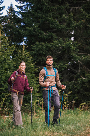 A young man and woman equipped with backpacks, hiking poles, and hiking clothes carefully navigate a rough mountain path surrounded by dense bushes.の写真素材