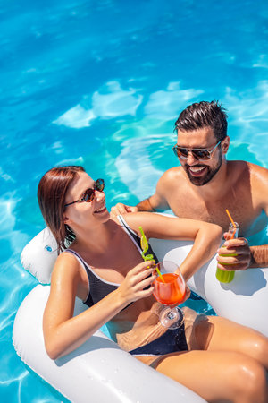 Young couple relaxing in the pool on a sunny summer day; the girl is on a floatie, the guy is beside...