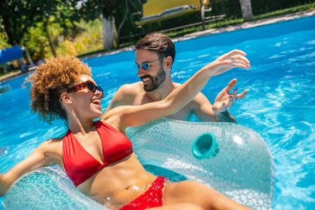Young couple enjoying a sunny summer day in the pool; the afro-haired girl is relaxing on a floatie while her partner is next to her, both sipping colorful cocktails and having fun.の写真素材
