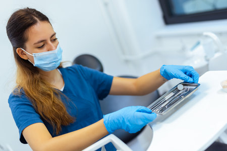 Female dentist wearing blue coat and face mask preparing dental tools in modern clinic. Concept of hygiene, readiness, and professional care.の写真素材