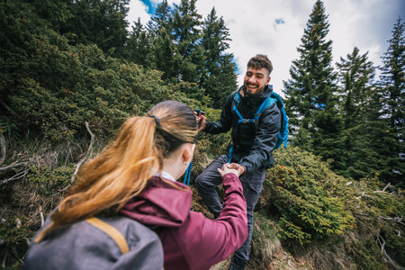 Hiking couple with full gear climbs a mountain trail, the man helping the woman by holding her hand, surrounded by breathtaking scenery.の写真素材