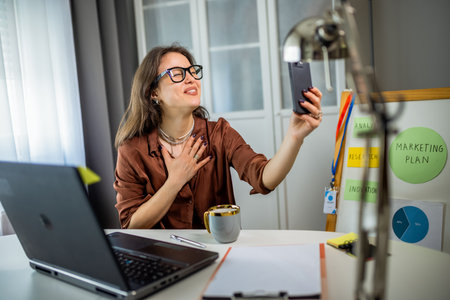 Young woman wearing glasses works from home on her laptop, presenting notes on a board and paper...