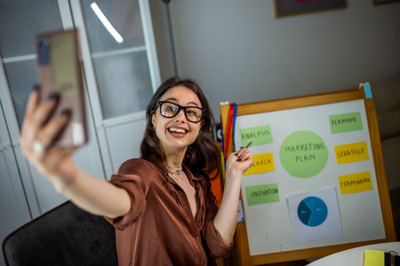 Young woman wearing glasses works from home on her laptop, presenting notes on a board and paper...