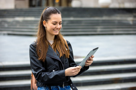 Young female student stands outside college campus holding a tablet, ready for studying and learning.の写真素材