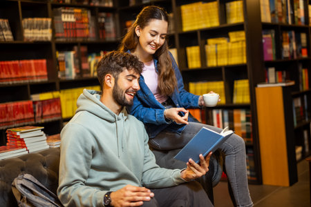 Young female and male students in a cozy library cafe, reading a book together while enjoying coffee, surrounded by shelves of books.の写真素材