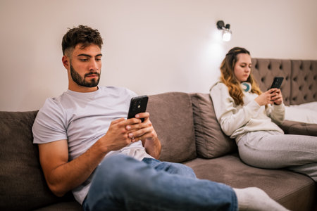 Young couple on vacation in a hotel room, sitting on the same couch, each on their own phone and headphones, absorbed in their own world.の写真素材