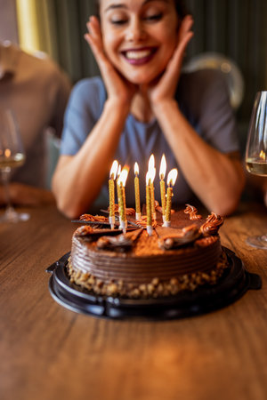 Birthday girl smiles as she blows out candles on her cake in a restaurant, surrounded by friends, gifts, and festive decorations.の写真素材