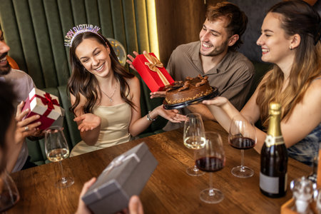 Friends celebrate a birthday at a restaurant, giving gifts to the birthday girl, sharing smiles, talking, and enjoying wine with cake on the table.の写真素材