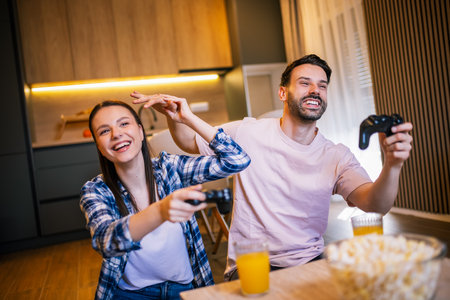Young couple in living room playing video games, teasing each other, popcorn and juice on table, laughing and enjoying fun time together.の写真素材
