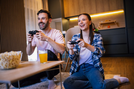 Happy young couple in living room playing video games with controller, smiling and enjoying time together, popcorn and juice on table in front.の写真素材