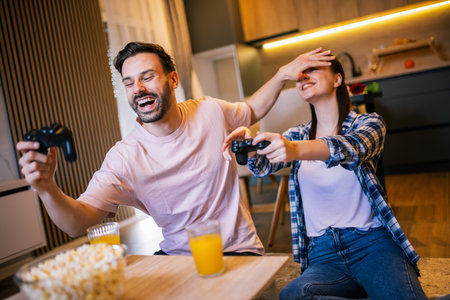 Young couple in living room playing video games, teasing each other, popcorn and juice on table, laughing and enjoying fun time together.の写真素材
