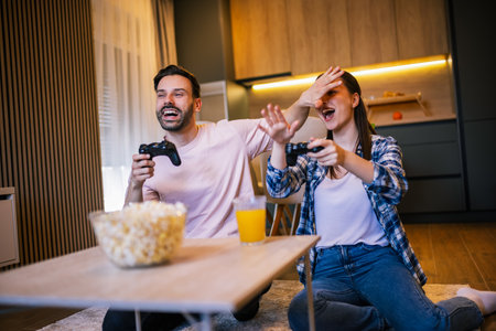 Young couple in living room playing video games, teasing each other, popcorn and juice on table, laughing and enjoying fun time together.の写真素材