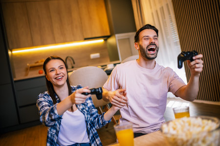 Young couple in living room playing video games, teasing each other, popcorn and juice on table, laughing and enjoying fun time together.の写真素材