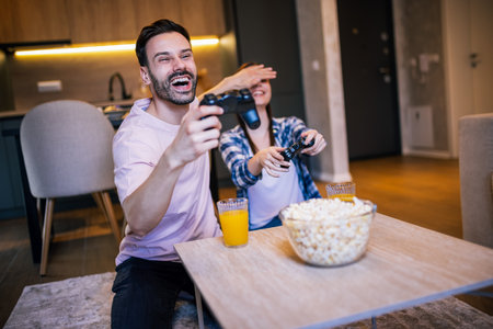 Young couple in living room playing video games, teasing each other, popcorn and juice on table, laughing and enjoying fun time together.の写真素材