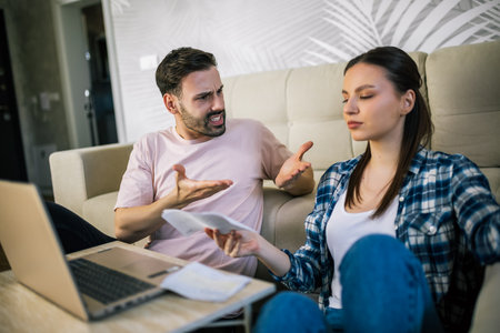 Young couple in living room sitting by laptop, holding unpaid bills, arguing over debt and financial stress.の写真素材