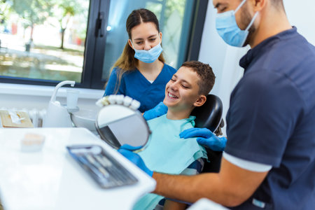 Little boy sits in dental chair, smiling happily as doctor and nurse hold a mirror to show his...