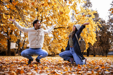 Happy young couple playfully throwing colorful autumn leaves in park, enjoying romantic fall day...