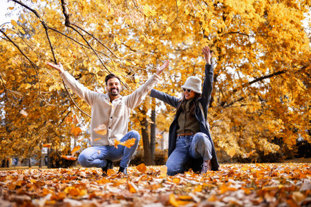 Happy young couple playfully throwing colorful autumn leaves in park, enjoying romantic fall day...