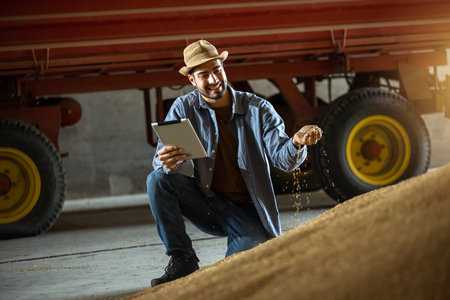 Young man holds soybeans in one hand and tablet in the other in warehouse, concept of modern agriculture, crop management, farming, logistics, food industry.の写真素材