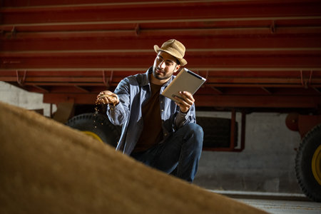 Young man holds soybeans in one hand and tablet in the other in warehouse, concept of modern agriculture, crop management, farming, logistics, food industry.の写真素材