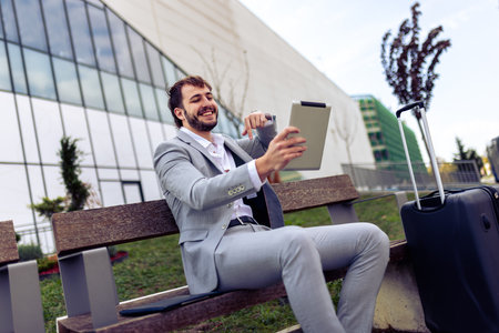 Professional businessman sitting on outdoor bench next to suitcase, conducting a video call on tablet, working remotely while on the go.の写真素材