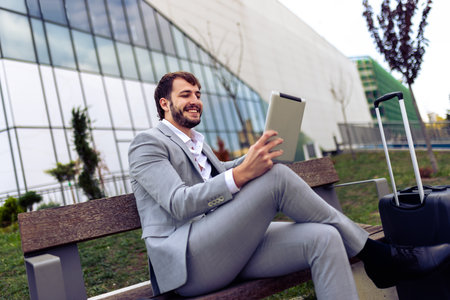 Professional businessman sitting on outdoor bench next to suitcase, conducting a video call on tablet, working remotely while on the go.の写真素材