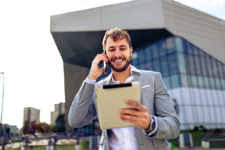 Confident businessman in suit standing outdoors in front of building talking on smartphone holding tablet working on the go.の写真素材