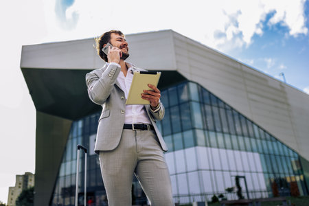 Confident businessman in suit standing outdoors in front of building talking on smartphone holding tablet working on the go.の写真素材