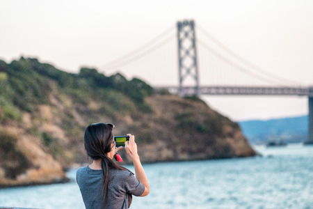 Female tourist photographing San Francisco Bay Bridge at sunset.