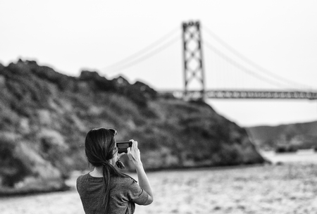 Female tourist photographing San Francisco Bay Bridge at sunset.の写真素材