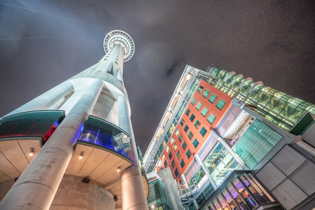 Auckland Sky Tower and surrounding buildings at night, skyward view from street level.