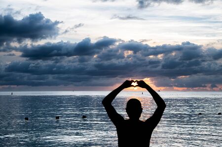 Man making heart sign with his hands with sunset on beackground. Holiday and relax concept.の写真素材