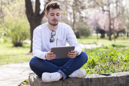 Handsome young man sitting in the park , use tablet and enjoy the outdoors. Selective focus and small depth of field.の写真素材